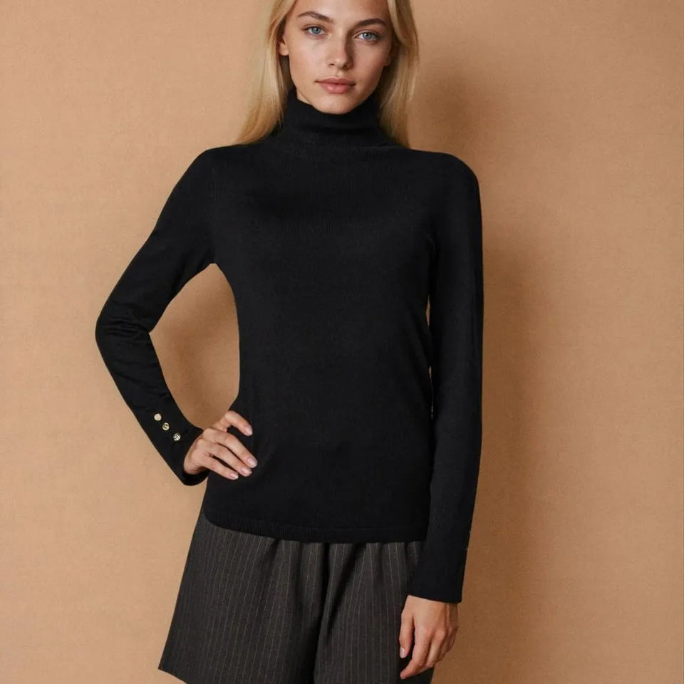 A woman modeling a black long-sleeve turtleneck sweater against a warm beige studio background. The sweater has three small gold buttons on the cuff and is paired with dark gray pinstriped trousers.