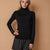 A woman modeling a black long-sleeve turtleneck sweater against a warm beige studio background. The sweater has three small gold buttons on the cuff and is paired with dark gray pinstriped trousers.