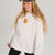 A young girl with long blonde hair wearing a light heather gray sweatshirt with a small embroidered teddy bear on the chest, paired with black pants and a dark cap.