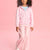 A young girl wearing a pink zip-up hoodie with white unicorn prints and matching pink pants, standing against a solid pink background.