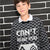 A boy wearing a black and white patterned knit sweater with the text 'CAN'T HEAR YOU' and a pixelated face graphic, layered over a white collared shirt, standing against a black brick wall.