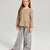 A young girl with long light brown hair stands against a light gray studio background, wearing a beige and brown leopard-print long-sleeve top with a peplum hem and wide-leg gray pants.