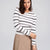 A woman wearing a fitted long-sleeve knit top with horizontal black stripes on a white background, paired with a beige skirt, against a light gray studio backdrop.