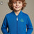 A smiling young boy with curly light brown hair wearing a bright blue zip-up jacket with a green and white stylized logo on the left chest, set against a solid warm gray background.