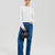 A model wearing a white ribbed knit long-sleeve top, blue jeans, and holding a small black leather handbag against a light gray studio background.