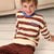 A smiling young boy wearing a brown and cream horizontally striped long-sleeve shirt with a blue collar and 'MONDAY AGAIN?' text on the chest, sitting on a light wood floor in front of a beige sofa.