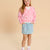 A young girl stands against a solid beige studio background wearing a pink cardigan with white daisy embroidery, a light blue denim skirt with pink bow details, a white t-shirt, white socks, and pink sneakers.