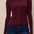 A close-up of a person wearing a ribbed long-sleeve cardigan in deep burgundy with a button-front closure, paired with blue jeans against a light textured wall.