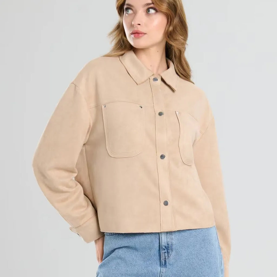 A woman wearing a cropped beige button-up shirt jacket with chest pockets, paired with light blue jeans, photographed against a light gray studio background.
