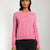 A woman wearing a pink long-sleeve button-up cardigan sweater and black pants, standing against a plain light gray studio background.