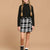 A young girl with long blonde braids is wearing a black ribbed mock-neck long-sleeve sweater and a black and white plaid mini skirt, standing against a solid beige studio background.