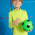 A smiling boy wearing a bright neon yellow-green long-sleeve athletic shirt holds a green soccer ball with black panels against a blue studio background with colorful geometric lines.