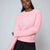 A woman wearing a pink ribbed knit sweater and black trousers, posed against a light gray studio background.
