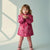 A young child wearing a pink hooded coat with a heart pattern, pink pants, and pink boots, standing against a pale blue-gray background with soft window shadows.