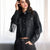 A woman wearing a black faux-leather cropped jacket with fringe details on the sleeves, paired with black jeans, posed in front of a bright window.