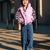 A young girl smiling outdoors while wearing a shiny pink puffer vest over a black-and-white striped long-sleeve top and blue jeans.