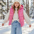 A young girl wearing a shiny pink puffer vest over a cream ribbed sweater, blue jeans, and a white knit beanie, standing in a snowy outdoor setting with trees in the background.