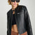 A close-up of a model wearing a black faux-leather moto jacket with a zippered front and a zippered chest pocket, styled with a black crop top and brown textured pants against a light gray background.