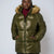 A woman wearing a shiny olive green puffer jacket with a tan faux-fur trimmed hood, layered over a burgundy turtleneck, against a plain light gray studio background.