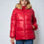 A woman wearing a shiny red puffer jacket with a tan faux-fur trimmed hood, paired with blue jeans, against a light gray studio background.