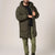 A man wearing a long hooded puffer parka in olive green, styled over a black shirt and olive green pants, standing against a plain light gray studio background.