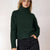 A woman wearing a dark forest green ribbed turtleneck sweater and black jeans, standing against a plain light background.
