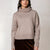 A woman wearing a light taupe knit turtleneck sweater and a dark brown velvet skirt, standing against a plain light gray studio background.