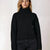A woman wearing a black ribbed turtleneck sweater and dark jeans, photographed against a plain light background.