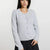A woman wearing a light gray knit cardigan sweater with a round neckline and button-front closure, paired with dark pants, against a plain light background.