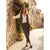 A woman stands on a sunlit cobblestone street wearing a white button-up shirt, a dark charcoal vest, dark brown cropped trousers, and dark loafers, leaning against a rustic stone wall with purple flowers and buildings in the background.