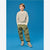 A young boy stands against a solid blue studio background wearing a beige and gray striped long-sleeve shirt, olive green cargo pants, and gray and beige sneakers.