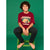 A smiling boy with curly dark hair sits barefoot on a green stool against a solid green studio background, wearing a burgundy graphic T-shirt with 'Adventure 20 CALL2026' and a tent illustration, paired with dark pants.