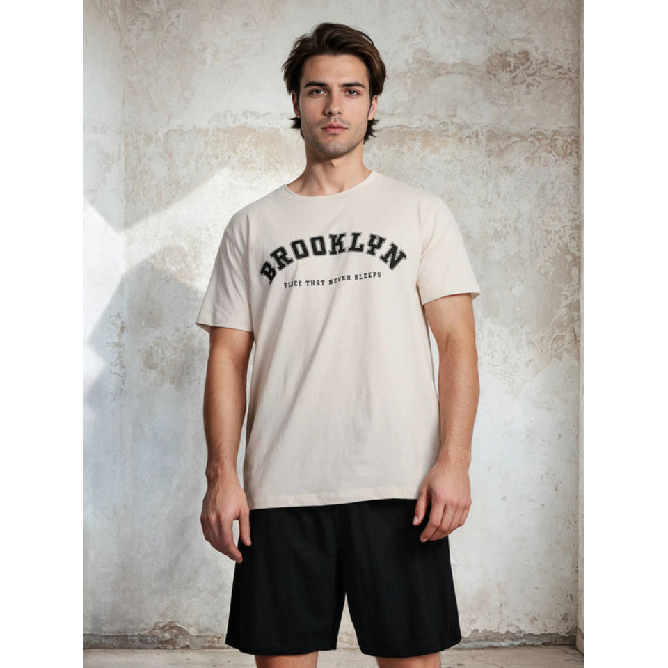 A man wearing a cream-colored short-sleeve t-shirt with 'BROOKLYN' printed in black arched text across the chest, paired with black shorts, standing against a textured concrete wall.