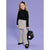 A young girl stands against a solid lavender studio background wearing a black ribbed mock-neck sweater, a gray and white striped mini skirt, and matching gray striped wide-leg pants. She is holding a small black backpack with pink pom-poms and 'HEART' printed on the front.