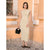 A woman stands outdoors in front of a peach-colored building, wearing a fitted sleeveless midi dress in a light beige or cream tone. She is holding a small black handbag and wearing black strappy flats.