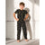 A young boy stands barefoot against a textured concrete wall, wearing a black short-sleeve pajama set with a yellow graphic pattern of cartoon characters.