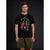 A man modeling a black crewneck t-shirt with a colorful graphic of a campfire scene and the text 'FOOLishly NORMAL CAMPING'. He is wearing olive green shorts against a dark studio background.