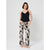A woman modeling a black camisole top and black wide-leg pants with a beige palm leaf print, standing against a light gray studio background.