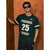A man wearing a dark green short-sleeve t-shirt with white graphic text and numbers, paired with black pants, posing against a textured orange-brown wooden wall.