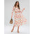 A woman modeling a long-sleeve midi dress with a pink, blue, and yellow floral print on a cream background, featuring a brown belt at the waist. She is holding a brown handbag and wearing beige strappy heels against a light gray studio background.