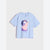 A light blue short-sleeve crewneck t-shirt featuring a colorful graphic print of a girl in a pink dress and purple headband, displayed against a light gray background.