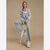 A woman models a light gray two-piece tracksuit consisting of a cropped zip-up jacket with white side stripes and matching wide-leg pants, sitting on a white stool against a solid beige studio background.