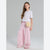 A young girl stands against a light gray studio background wearing a white short-sleeve top with small dot details, pink and white vertically striped wide-leg pants, and a round beige woven crossbody bag.