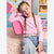 A young girl with braided hair sits on a pink shag rug wearing a light pink long-sleeve top with tiny darker pink heart-like dots, light blue jeans, and a pink belt with a silver buckle. She is posed in a colorful children's room setting.