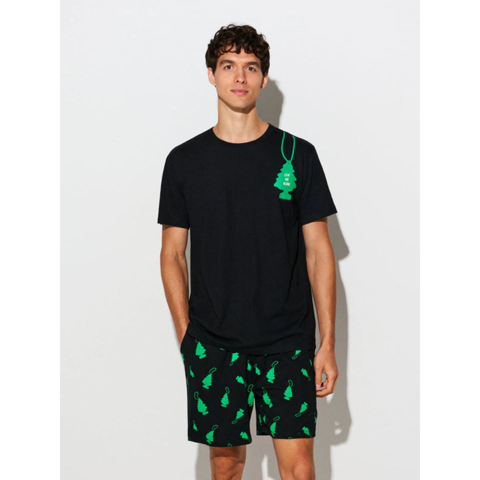 A man modeling a black short-sleeve t-shirt with a green tree-shaped hanging ornament graphic on the chest, paired with black shorts featuring a green tree pattern, set against a plain light gray background.