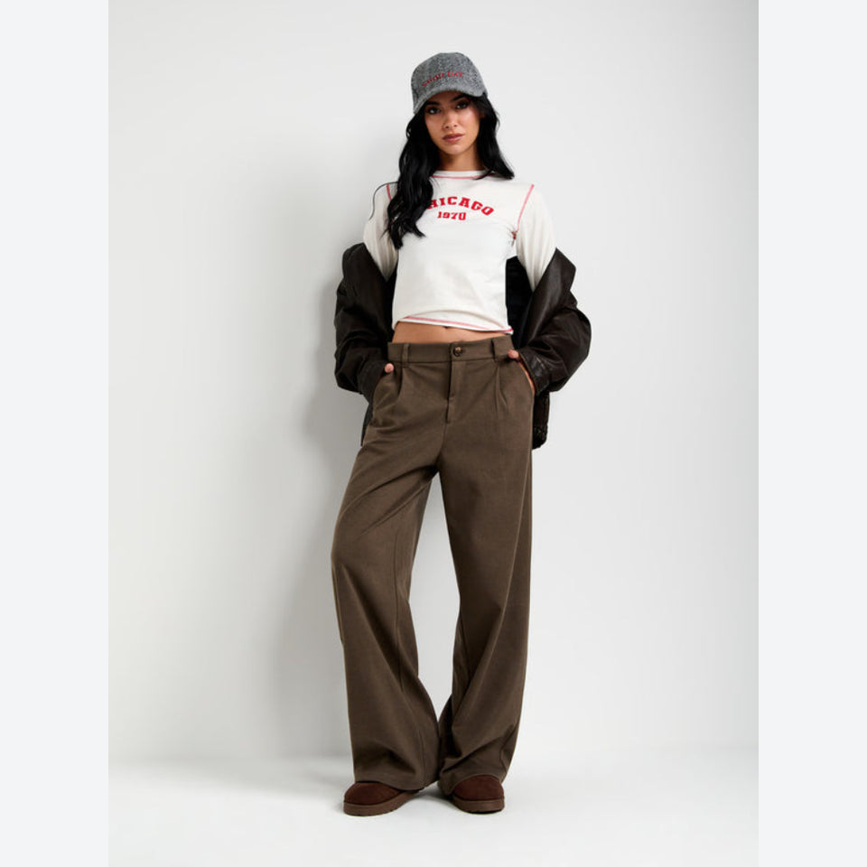 A model stands against a light gray studio background wearing a white cropped sweatshirt with red text, a black-and-brown jacket draped off the shoulders, brown wide-leg pants, and a gray cap.