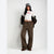 A model stands against a light gray studio background wearing a white cropped sweatshirt with red text, a black-and-brown jacket draped off the shoulders, brown wide-leg pants, and a gray cap.