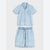 A light blue denim two-piece set for a child, featuring a short-sleeve button-up shirt with a chest pocket and matching drawstring shorts, laid flat against a light gray background.