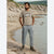 A man stands on a rocky beach shoreline wearing a beige graphic t-shirt, light gray cargo jogger pants, and brown sneakers, with water and rocky cliffs in the background.
