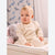 A baby sitting on a changing table wearing a cream ribbed long-sleeve top and matching cream pants, with a gray stuffed elephant toy beside them and a patterned curtain in the background.